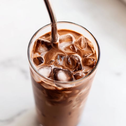 A clear glass holds the homemade Milo Ice Latte Hack, with ice cubes and a spoon stirring the chocolatey mixture.  