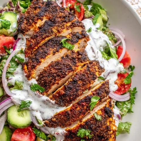 Sliced spiced grilled chicken rests atop crisp cucumbers, tomatoes, and greens, drizzled with creamy yogurt dressing on a plate.  