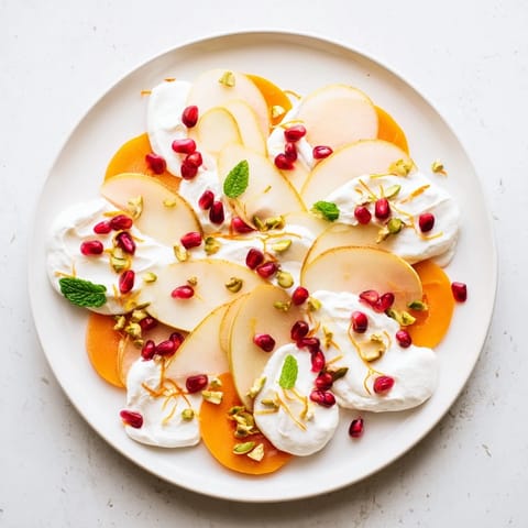 A festive persimmon and pear snowscape featuring vibrant pomegranate seeds and chopped pistachios on a white plate.