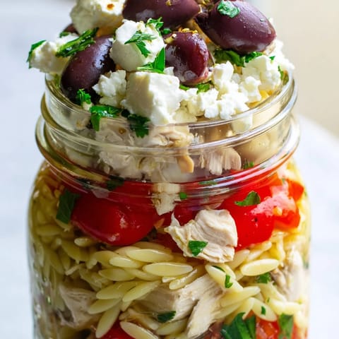 Close-up view of colorful Mediterranean chicken salad in a jar with Kalamata olives, red onion, and fresh parsley on top.